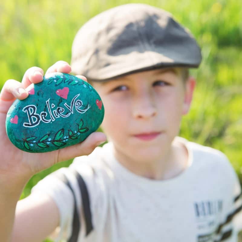 Image of boy with cap holding rockwith the word 'Believe' painted on it.